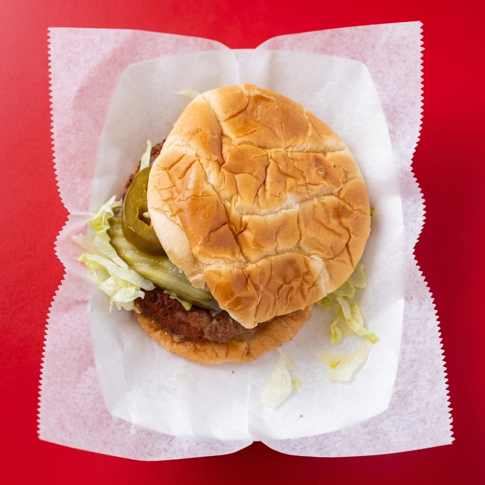 A hamburger with lettuce, pickles, and jalapeños in a paper wrapper on a red surface.